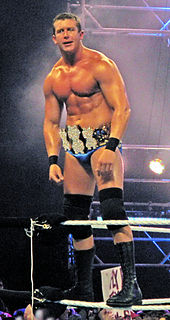 A dark-haired man wearing blue wrestling tights, black knee-high wrestling boots and black knee-pads stands on the white ropes of a wrestling ring. He is wearing around his waist a gold-plated professional wrestling championship belt shaped with three dollar signs on the front.