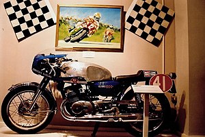 Race prepared road bike with race number 41, a large shiny aluminium fuel tank and streamlined top half fairing with dropped handlebars in front of a wall mounted large painting of Mike Hailwood in 1978 on a TT race prepared Ducati leading Phil Read on a Honda flanked by twin chequered flags