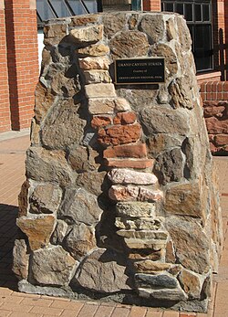 Um pilar robusto de pedra motorizada de forma irregular com inserções de pedra em forma de tijolo empilhadas formando uma coluna inclinada para a direita.  Uma placa no pilar diz: "Grand Canyon Strata, cortesia do Grand Canyon National Park".