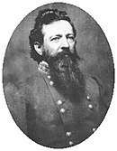 Blank and white photo shows a black-haired man with a heavy beard. He wears a gray military uniform with two rows of buttons.