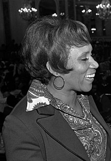 A smiling African-American woman, facing left. Her hair is cut in bangs, and she is wearing a high-collared blouse.