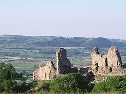 In the foreground a ruined castle built of silvery-grey stone; in the background a landscape of rolling hills, with fields bounded by hedges and small groups of trees.