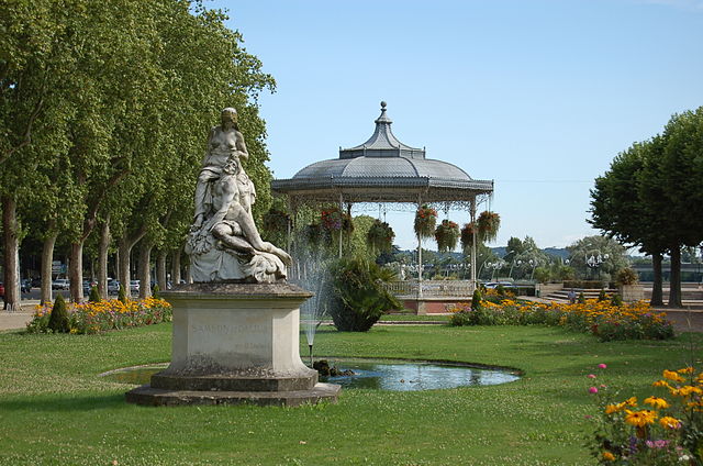 Photo représentant la sculpture d'un homme dévêtu aux pieds d'une femme seins nus, devant un kiosque à musique, dans un parc arboré.