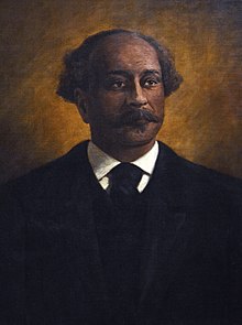 Retrato pintado hasta el busto de un hombre negro con bigote y un ligero mechón de vello facial en el labio inferior, vestido con traje negro y corbata sobre un fondo dorado iluminado.  Su cabeza es calva en la parte superior con mechones de cabello rizado grisáceo que sobresalen a ambos lados de la cabeza detrás de las sienes.