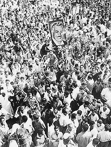 A crowd of people, many waving. One person is holding up a portrait of a man