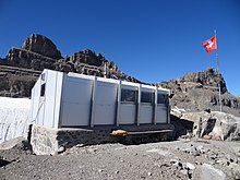 Une cabane métallique blanche en premier-plan, un mat avec un drapeau suisse à sa droite et des montagnes rocheuses en arrière-plan.