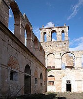 Fotografia scattata tra le rovine di un edificio in pietra, che mostra terra al posto del pavimento e muri di pietra grezza che si elevano a tre piani con finestre e cornici mancanti.  I ruderi non hanno tetto e non hanno soppalchi di livelli superiori.