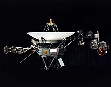 A space probe resting on a stand, with a parabolic antenna pointing upwards and two arms extending from the sides, bearing cameras and other devices, against a black background curtain