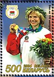 Eine blonde Frau in einer weiß-roten Jacke hält mit der linken Hand eine Goldmedaille hoch.