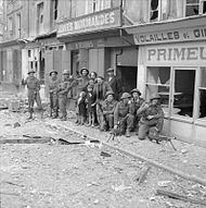 A mixed group of soldiers and civilians in front of some damaged shops; the street is littered with debris