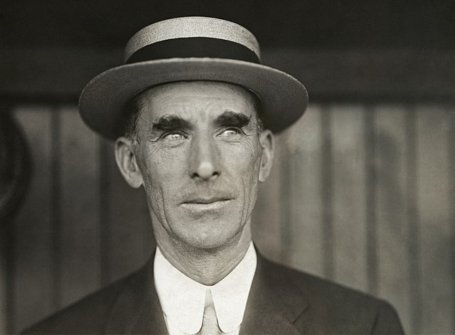 A man in a straw boater with a dark band and a dark suit, white shirt, and light-colored tie (black-and-white photo from the shoulders up)