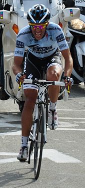 A road racing cyclist in a blue and black jersey with white trim pedaling hard, with a grimace on his face. A motorcycle follows behind.