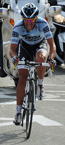 pelumba basikal jalan lumba dalam birudan jersi hitam dengan putih memangkas mengayuh keras, dengan Grimace pada mukanya. Motorsikal berikut belakang.