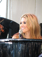 Een blonde vrouw lacht terwijl ze naar rechts kijkt terwijl ze achter een piano zit