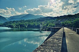 Immagine del lato nord-ovest della diga fotografata dallo sbarramento in un pomeriggio estivo lievemente ventoso. Cielo azzurro con nuvole bianche.