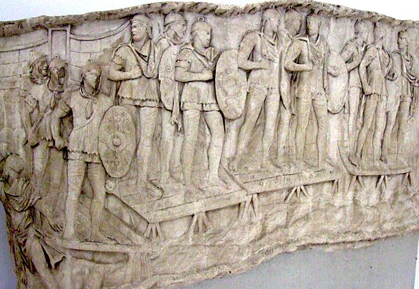 Roman auxiliary infantry crossing a river, probably the Danube, on a pontoon bridge during the emperor Trajan's Dacian Wars (AD 101–106). They can be distinguished by the oval shield (clipeus) they were equipped with, in contrast to the rectangular scutum carried by legionaries. Panel from Trajan's Column, Rome