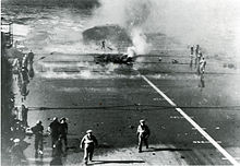 The wreckage of the aft lift is visible through the smoke behind the hole made by the only bomb to penetrate through the ship's flight-deck armour IllustriousFlightDeck10Jan41.jpg