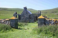 A grass-covered driveway leads between two stone pillars with orange lichen on their pyramid-shaped tops. Beyond is a stone building with ruined outhouses and green hills with a blue sky in the background