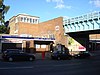 Um edifício de tijolos marrons com uma placa retangular azul escura dizendo "RUISLIP MANOR STATION" em letras brancas, tudo sob um céu azul claro