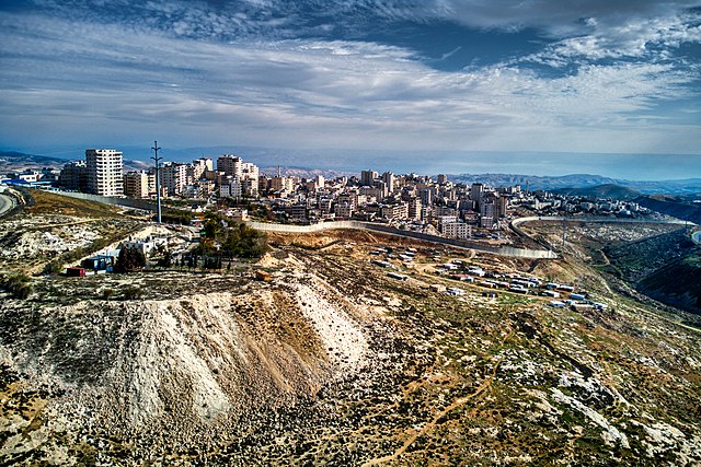 גדר ההפרדה מתפתלת ומקיפה את מחנה הפליטים שועפאט ואת ענאתא. לפני הגדר ניתן לראות מבנים של בדואים ובמרחק את מדבר יהודה, בקעת הירדן ועבר הירדן.