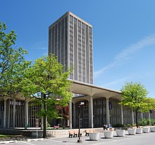 Een enkele toren in moderne stijl is omgeven door een lager openluchtpaviljoen met bomen die het gebied accentueren.