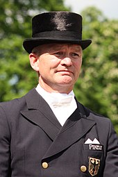 Das Foto zeigt das Gesicht des Dressurreiter Hubertus Schmidt. Er trägt einen schwarzen Zylinder und einen schwarzen Frack.