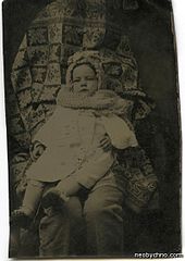 Toddler in a white coat and knitted hat and scarf sat on a pair of trousered knees. The head, torso and arms of the second person are hidden under fabric.