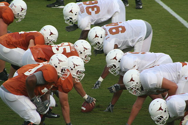 The offensive line (on left, in orange jerseys) consists of a center (with ball in hand ready to snap) with two guards on either side, and two tackles