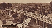 Photo en noir et blanc du peloton du Tour de France passant sur un pont au-dessus d'une rivière.