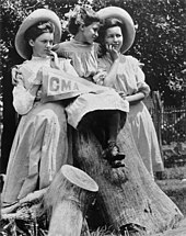 Photograph of three female students wearing dresses sitting on a tree stump, one of whom is holding a pennant readings "CMA"