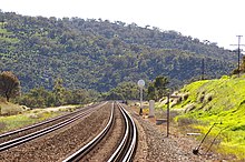 Two parallel dual gauge railway tracks in a rural area with a large hill in the background