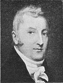 Black and white image of a male with receding hair wearing a dark suit and cravat
