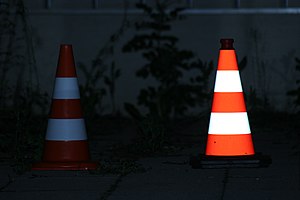 Links ein eingefärbter Leitkegel mit (schwach) retroreflektierenden weißen Streifen im Scheinwerferlicht, rechts ein voll-retroreflektierender Leitkegel nach TL