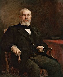 Bir koltukta oturan bıyıklı sakallı bir adamın resmi.