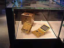 Sous une vitrine, on observe deux consoles de jeu dorées et logotées.