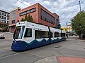 Straßenkreuzung mit zweigleisiger Straßenbahnstrecke und dreiteiligem Straßenbahn-Triebwagen in Bildmitte. Den Hintergrund dominiert ein in dunklen, teils schmutzigen Rottönen gehaltenes modernes viergeschossiges Geschäftsgebäude. Der Triebwagen ist weiß und hat im unteren Viertel auf ganzer Länge ein stilisiertes Meereswellen-Muster in drei dunklen Blau- und Grüntönen. Die festen Anschriften weisen in Verbindung mit Tacoma-Link-Logos auf die Marke TACOMA LINK und den Betreiber SOUND TRANSIT hin. Auf dem Zielschild steht ST JOSEPH.