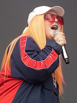 A woman is singing into a hand-held microphone with her eyes partly closed. The back of her right hand has an obscured image or a tattoo. She wears a white cap and rose-coloured glasses. Her hair is strawberry blonde and hangs down past her shoulders. She wears a red and blue jacket.