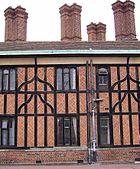 A close-up photograph of a building made with black timbers and red brick. The building has four tall, brick chimneys. A relatively modern drainpipe comes down the middle of the building.