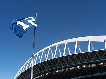 A blue flag with a white number 12 flies against a clear sky. An expansive white roof truss is behind the flagpole.