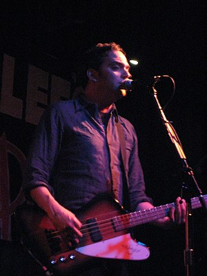 Adam Schlesinger is playing a guitar and singing live in front of a microphone.