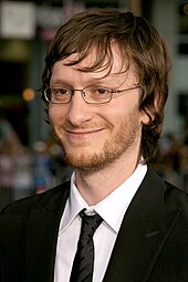 Una foto de perfil de un hombre rubio que sonríe.  Lleva gafas y esmoquin.