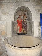 Photographie d’un ensemble statuaire polychrome dans une niche en pierre, devant une vasque de pierre également.