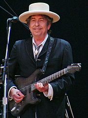 A gray-haired man wearing a hat plays a guitar.