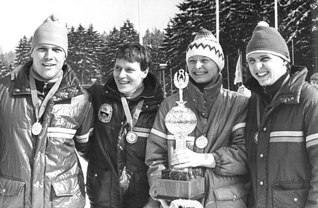 Four smiling people carrying medals around their neck are aligned shoulder-to-shoulder in an outdoor location with pine trees in the background. On the left, a man wears a shiny jacket and winter cap. Next to him, a second man with short dark hair wears a dark jacket with a badge. The third person is a woman wearing a jacket and an embroidered winter cap, and holds a trophy in her hands. The last person, on the right, is another man, also wearing a jacket and a winter cap.