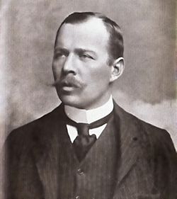 Head and shoulders portrait of a man with receding hair, heavy moustache, looking left from the image. He wears a high white collar, black necktie, dark waistcoat and jacket.