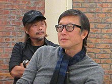 Lau is seated on the right, wearing a gray shirt and a blue striped scarf