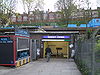 Un bâtiment en briques brunes avec un panneau bleu rectangulaire indiquant « HIGHGATE STATION » en lettres blanches et une personne marchant sous un ciel blanc