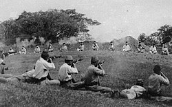 A dozen prisoners sit bound at a distance as Japanese soldiers in the foreground kill them