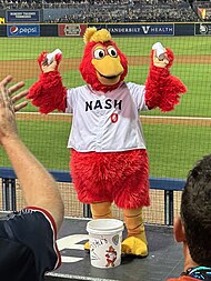 A person wearing a red anthropomorphized rooster costume dressed in a white baseball jersey with "NASH" across the chest in navy blue and a red "0" below it dances on a baseball dugout.