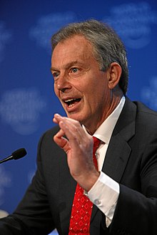 A photograph of a man with greying hair speaking into a microphone and gesturing with his left hand